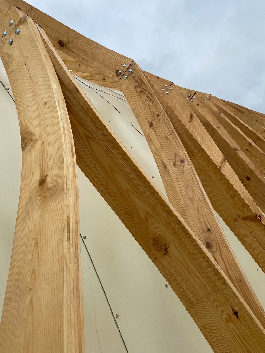Modern wooden roof truss curves upwards against grey sky.